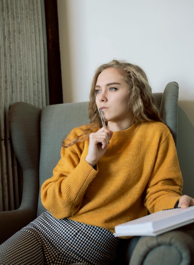 Woman in a mustard sweater sitting in an armchair, pen in hand, lost in thought.