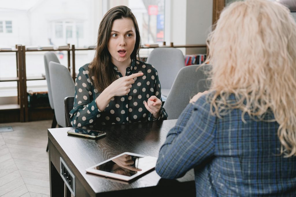 Astonished female pointing away while sitting at table with smartphone and tablet and talking to woman